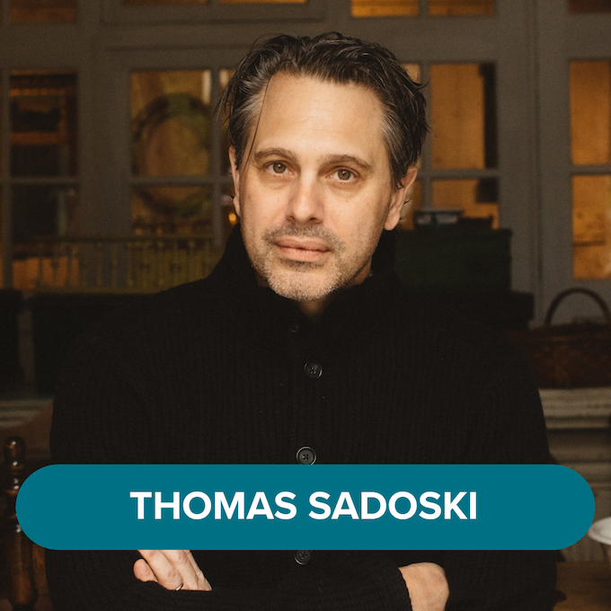 A man with dark hair and a beard, wearing a black sweater, sitting indoors with his arms crossed, in front of a background with windows and warm lighting. The text "Thomas Sadoski" is displayed at the bottom.
