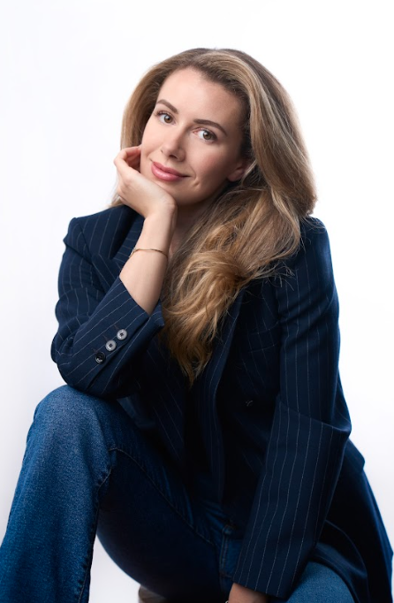 A woman with long, wavy light brown hair sitting against a plain white background. She is wearing a dark blue pinstripe blazer and blue jeans, with a slight smile and her chin resting on her hand.