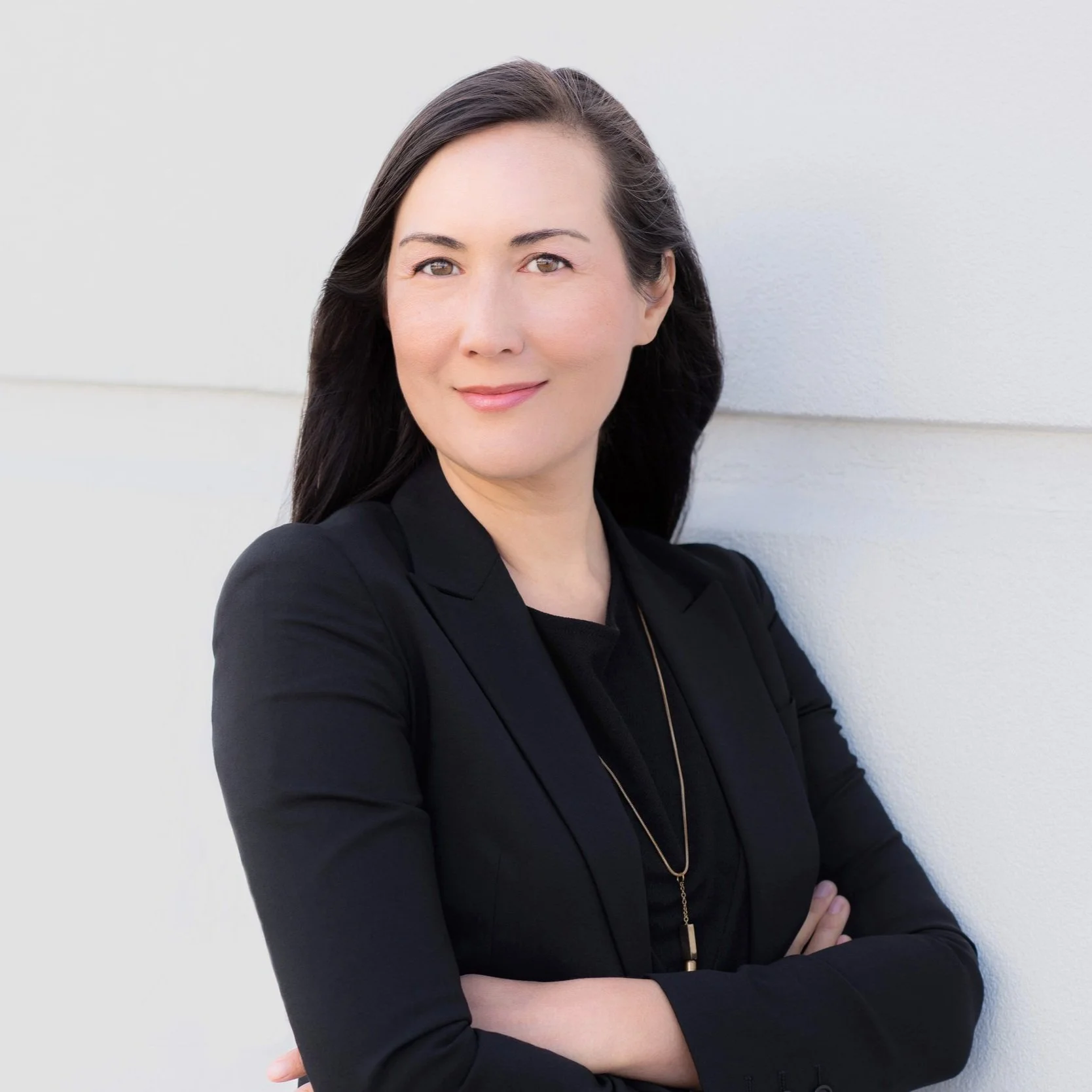 Portrait of a woman with long dark hair wearing a black blazer and standing against a white wall, crossing her arms and smiling.