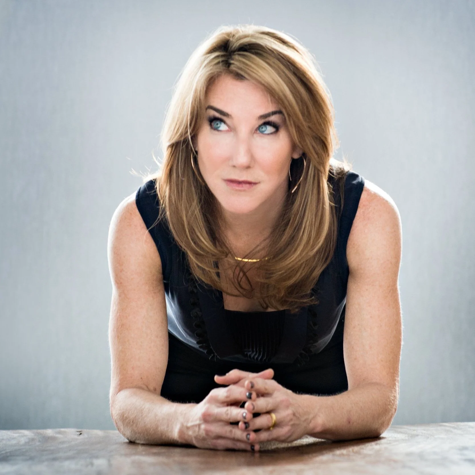 A woman with long reddish-brown hair and blue eyes, wearing a sleeveless black top, resting her arms on a table and looking to the side against a plain gray background.