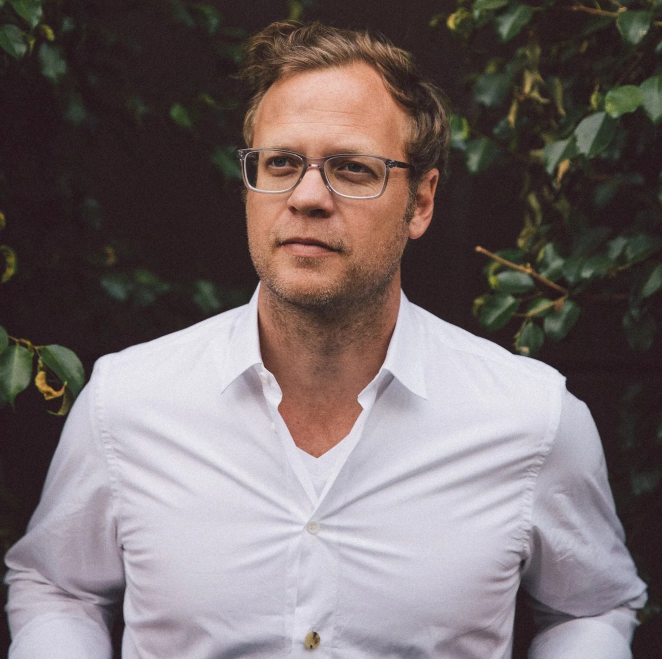 A man with light brown hair, glasses, and a short beard, wearing a white button-up shirt, standing outdoors in front of green leafy plants.