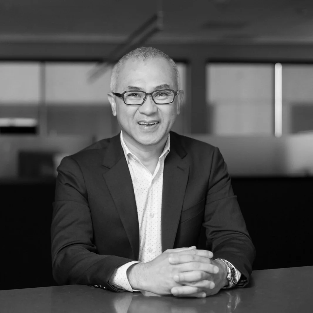 Black and white portrait of a man with glasses, short hair, wearing a suit and white shirt, sitting at a table with hands clasped, smiling at the camera in an office setting.