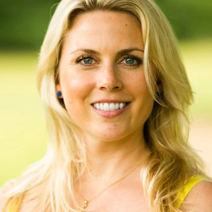 Close-up of a smiling blonde woman with blue eyes, wearing a yellow top and a gold necklace, outdoors with blurred greenery background.
