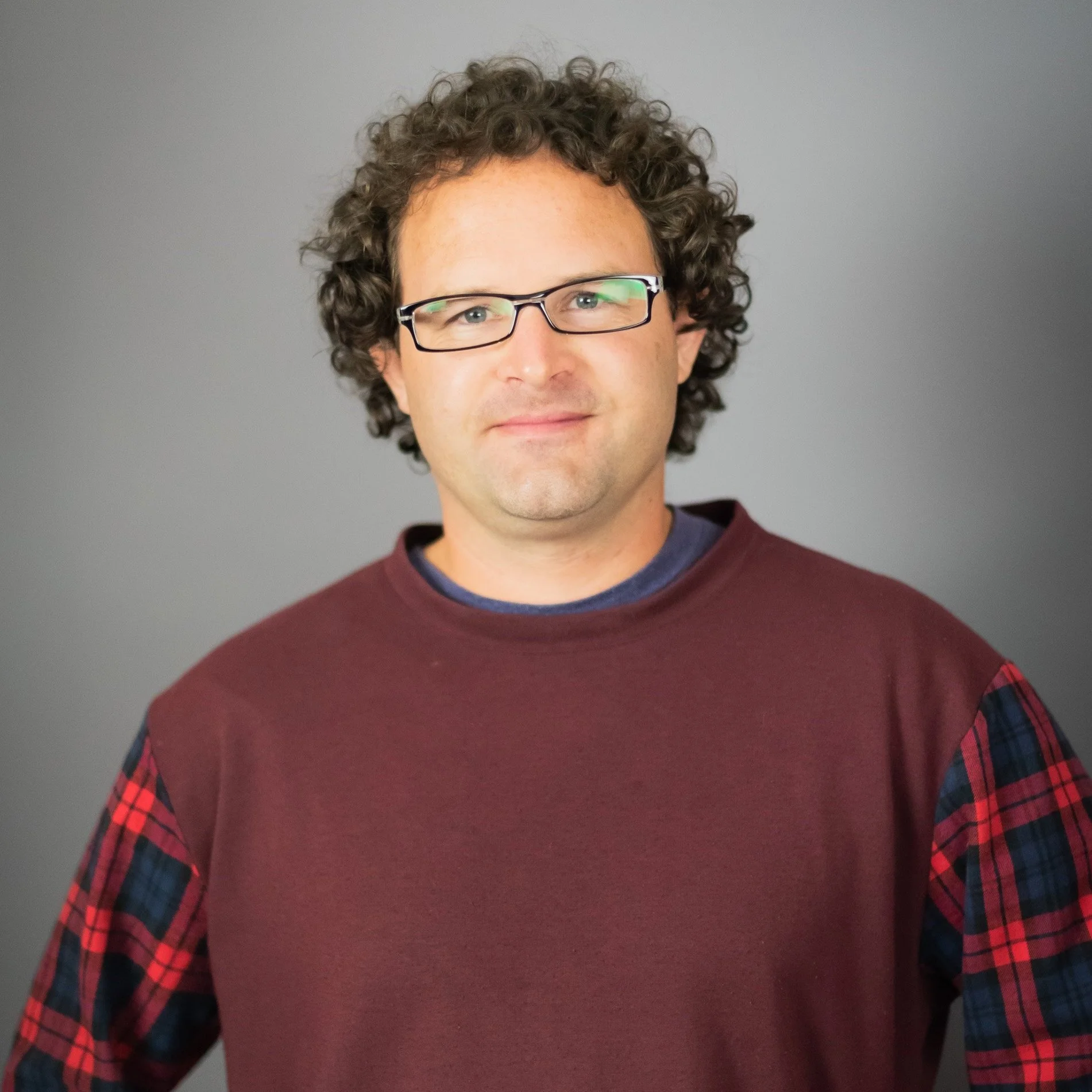 A man with curly brown hair, glasses, and a slight smile, wearing a maroon sweater with plaid pattern on the sleeves, standing against a gray background.