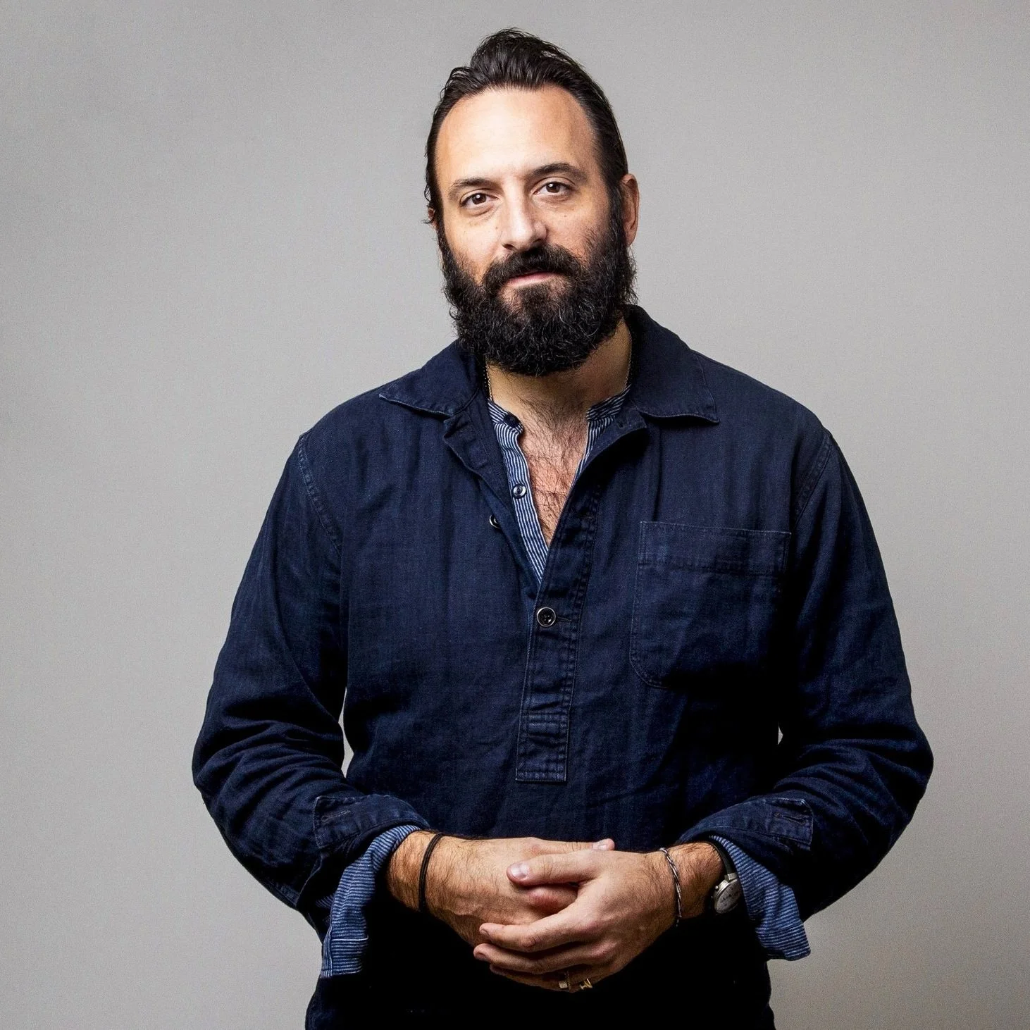 A man with a beard and dark hair wearing a dark blue button-up shirt standing against a light grey background.