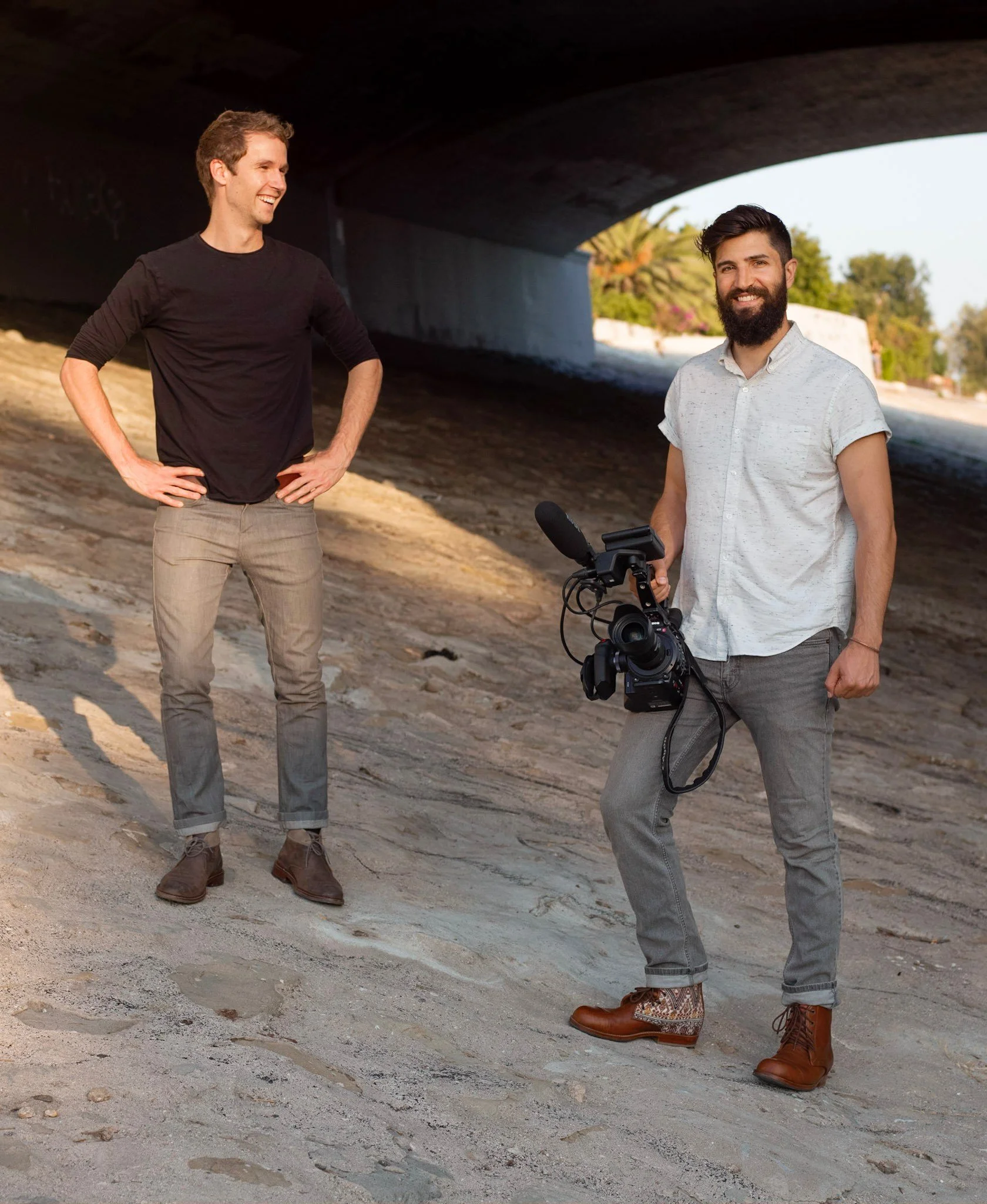 Two men standing outside under a bridge; one is holding a professional video camera, and both are smiling.