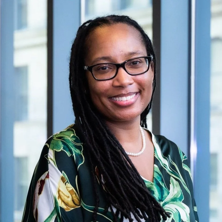 A woman with glasses, smiling, wearing a pearl necklace and a patterned green, white, and black top, standing in front of a window with a cityscape in the background.