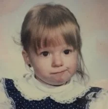 A young girl with brown hair and bangs, wearing a dark blue dress with white lace collar.