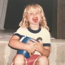 A young girl with blonde curly hair laughing and holding a toy or book, sitting outdoors near stairs.