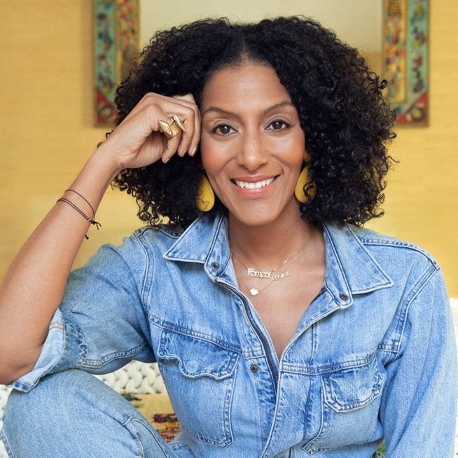 A woman with curly hair wearing a denim jacket and colorful earrings, smiling at the camera, sitting indoors with yellow walls and framed art in the background.