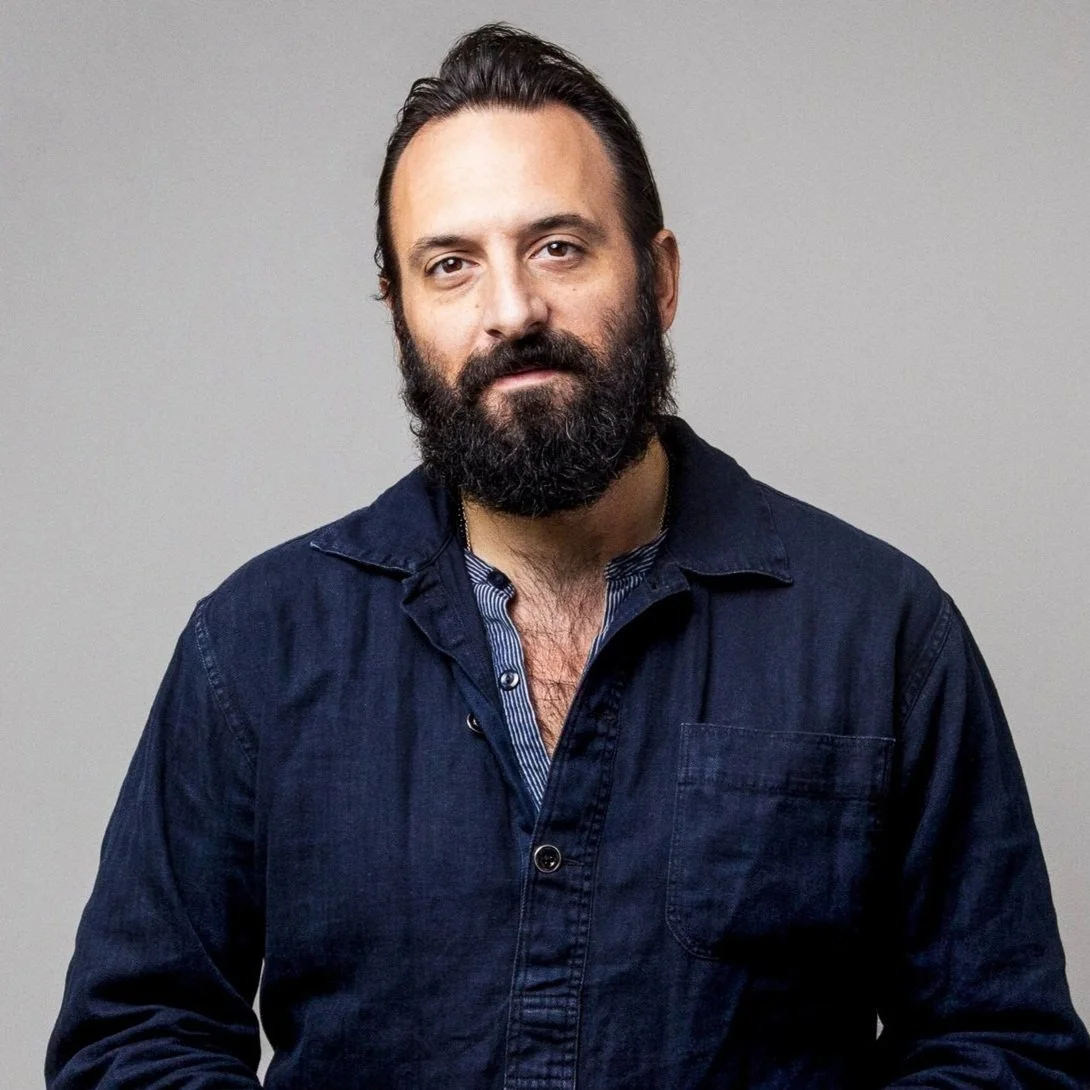 A man with a dark beard and hair, wearing a dark button-up shirt, looking at the camera against a plain gray background.
