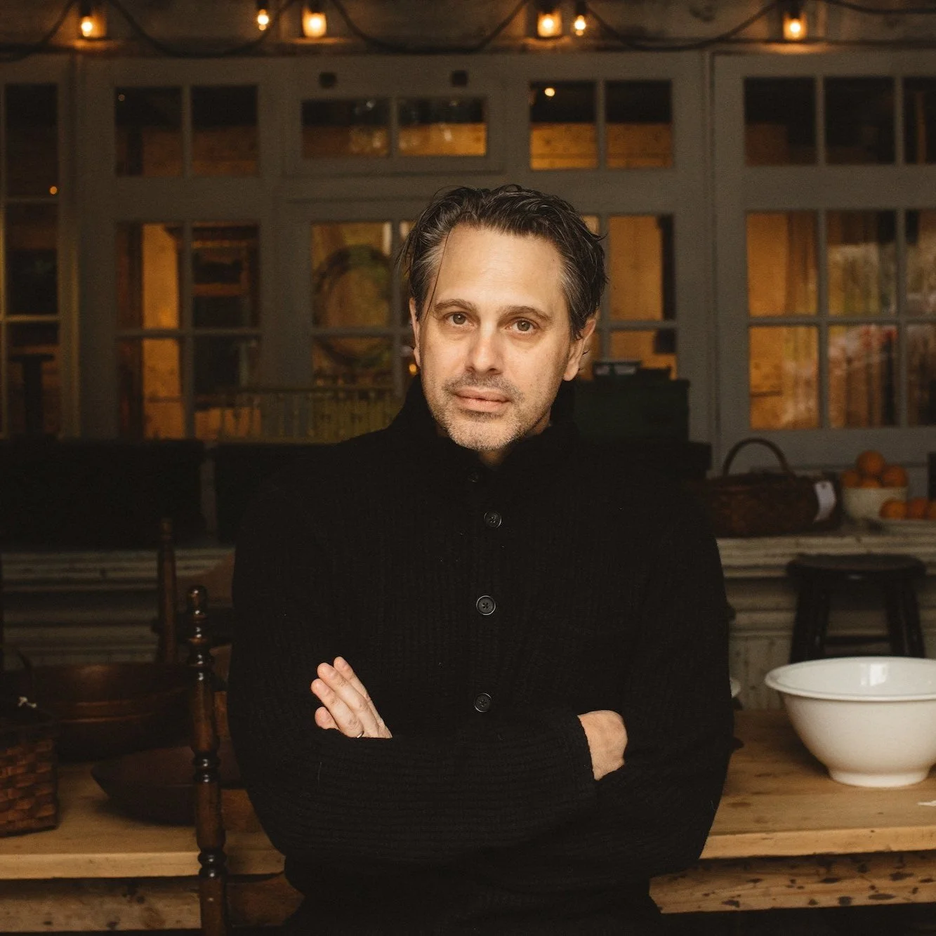 A man with gray hair and stubble wearing a black sweater, sitting with crossed arms in a warmly lit kitchen or dining area with wooden furniture and a large white bowl on the table.