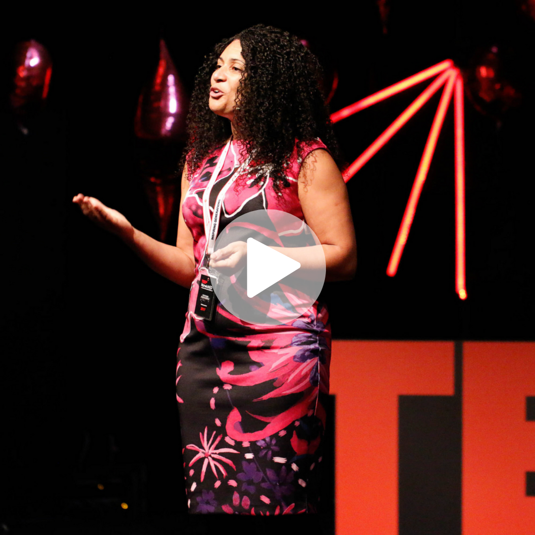A woman with curly hair giving a presentation on stage, wearing a colorful floral dress, with a black background and red neon lights.