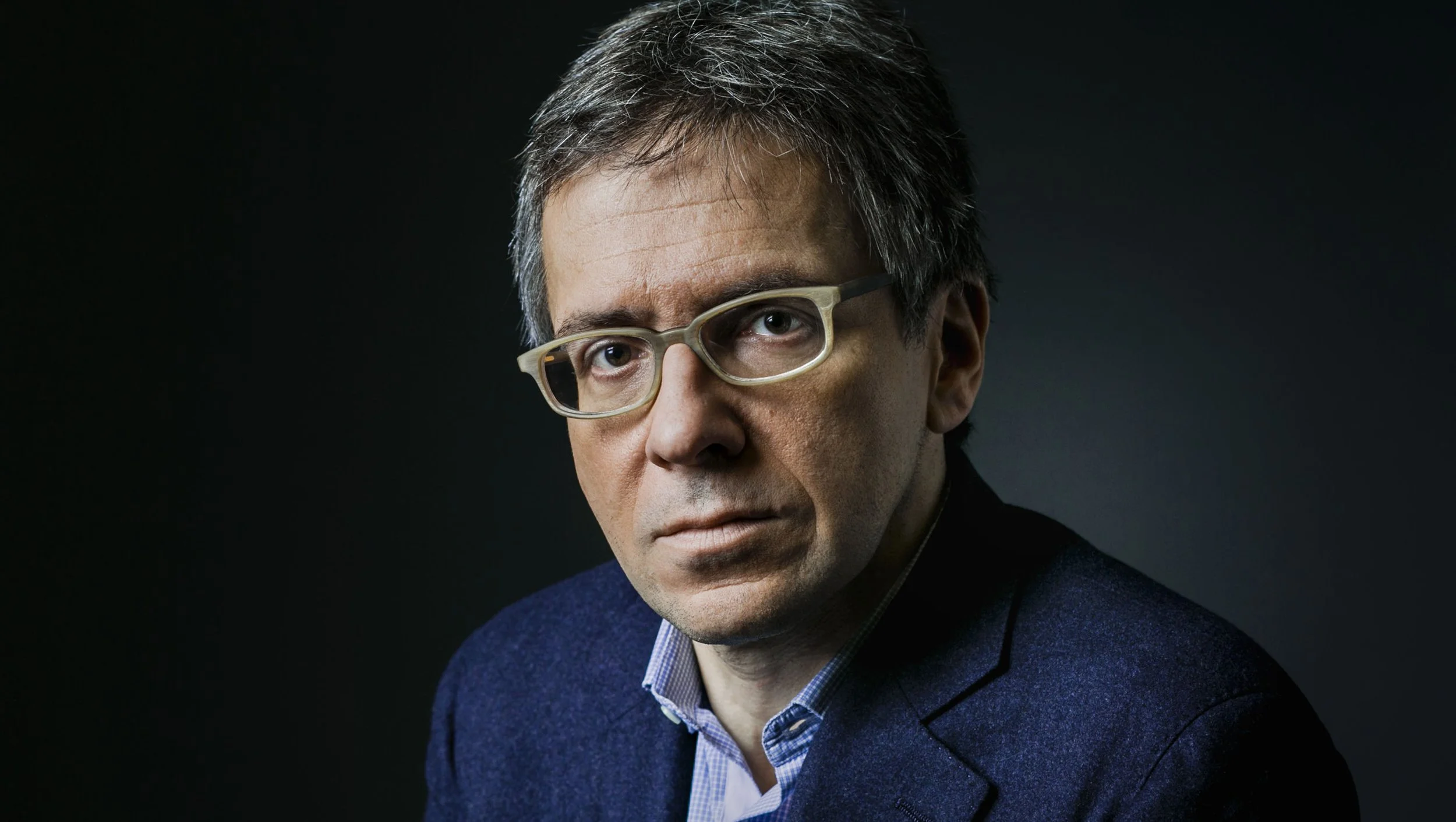 Portrait of a middle-aged man with glasses, wearing a blue blazer and a striped shirt, against a dark background.