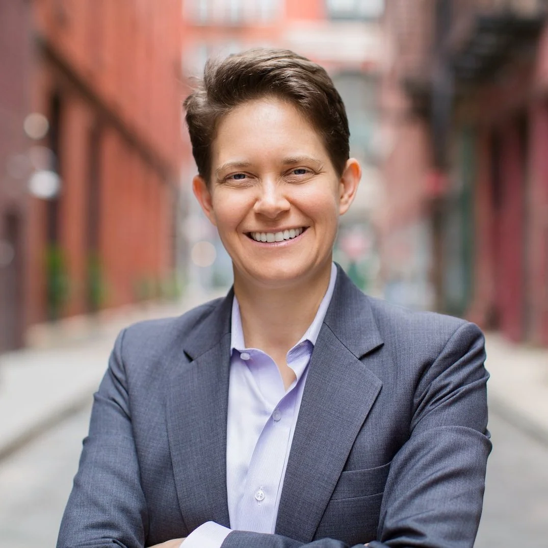 A smiling woman with short dark hair, wearing a gray blazer and a light purple shirt, standing in an urban alleyway with brick buildings in the background.
