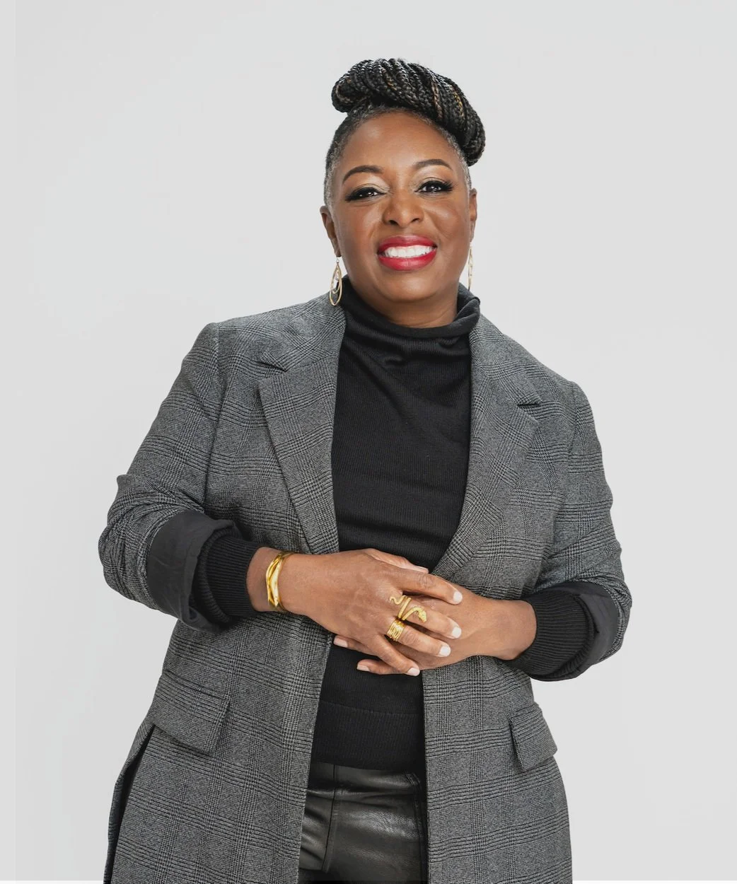 A confident woman with braided hair, wearing a gray plaid blazer over a black turtleneck, accessorized with gold jewelry, smiling against a plain light background.