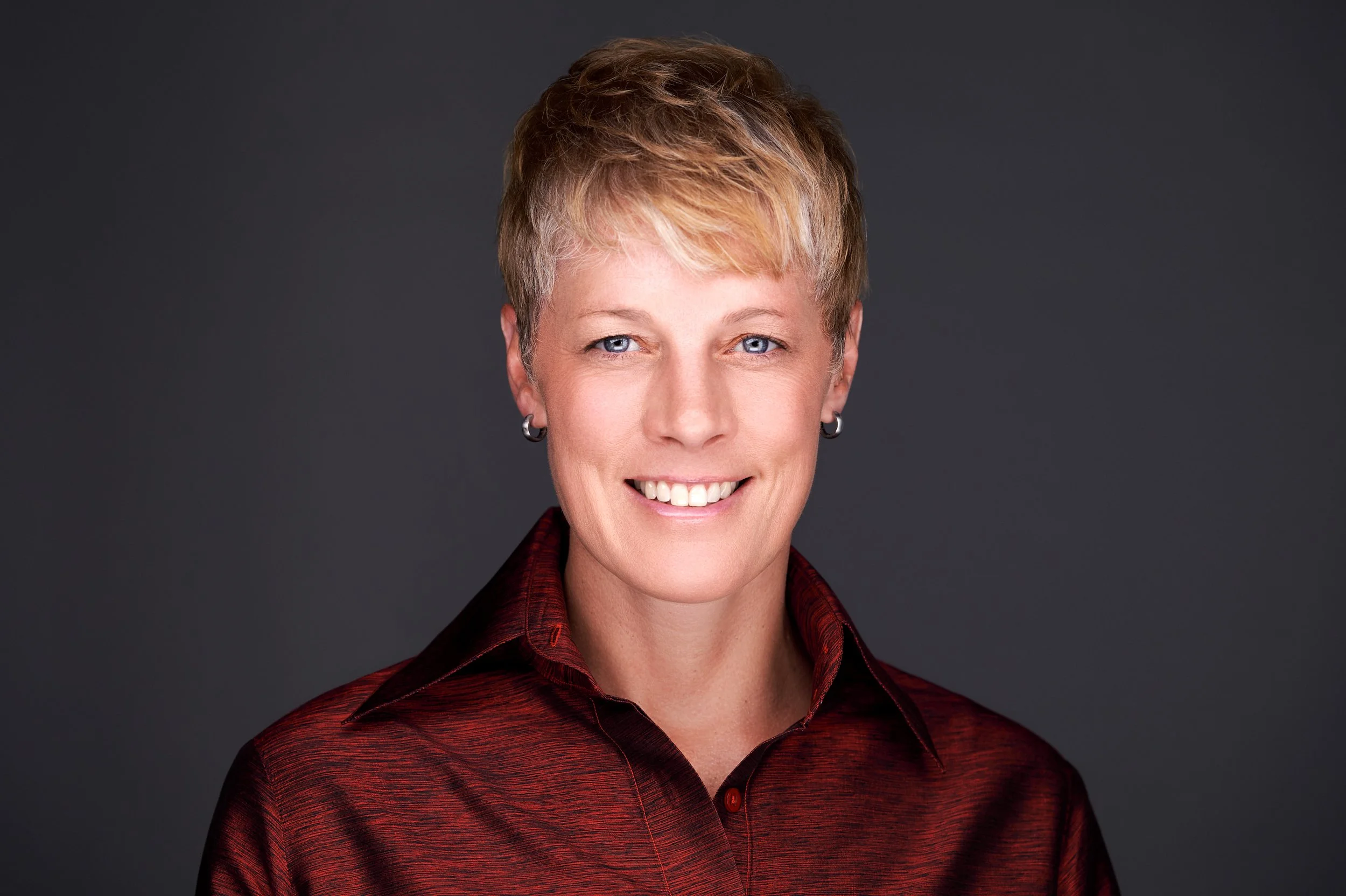 Portrait of a woman with short blond hair, blue eyes, wearing a red shirt and earrings, smiling against a dark background.
