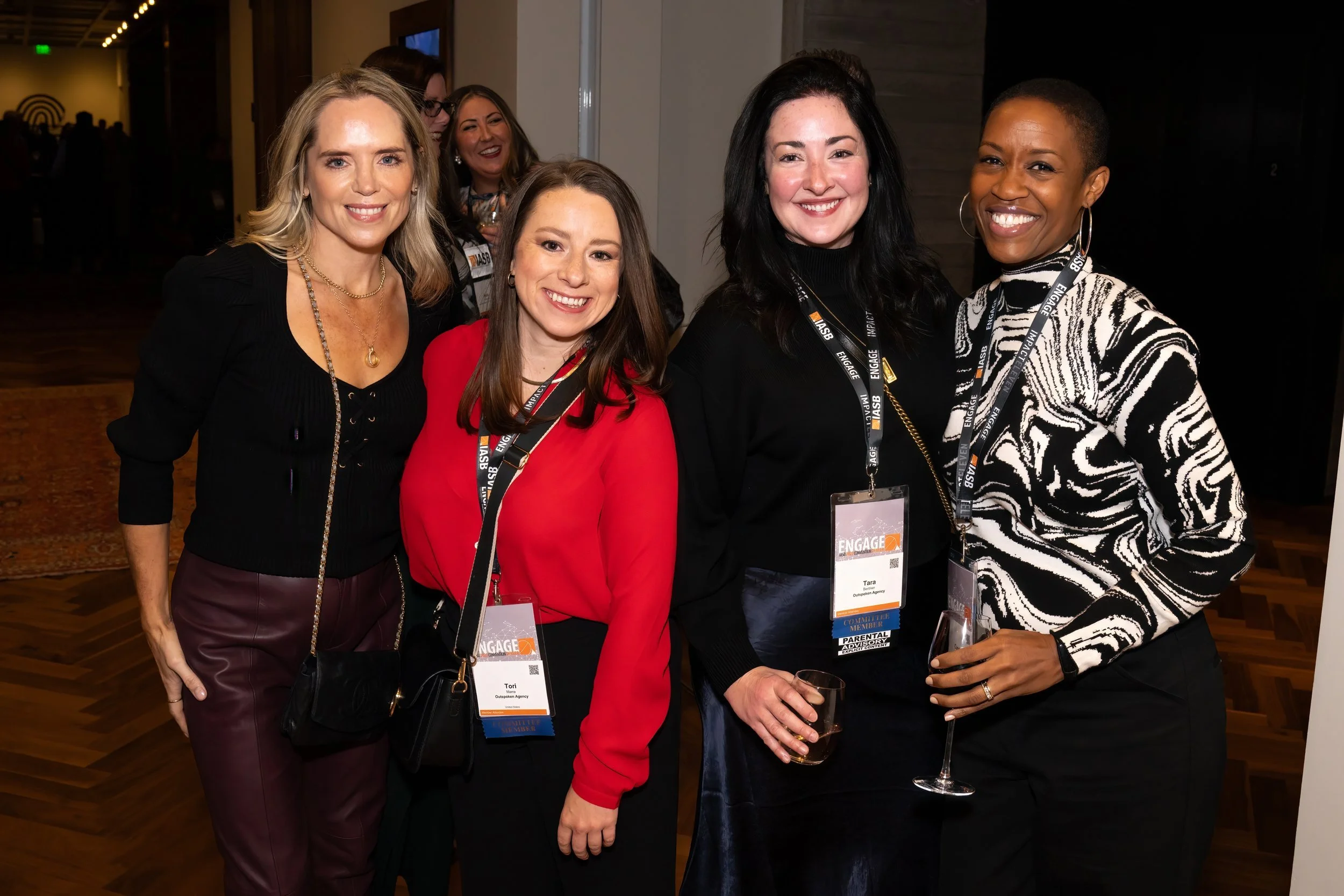 Group of four women smiling at an event, wearing conference badges, standing in a room with wood flooring and dark walls, holding drinks.