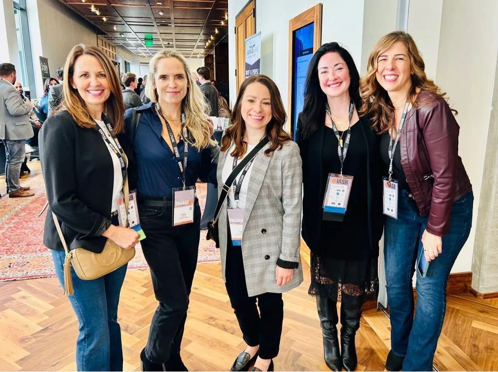 Five women standing close together at a conference, smiling and wearing name badges.