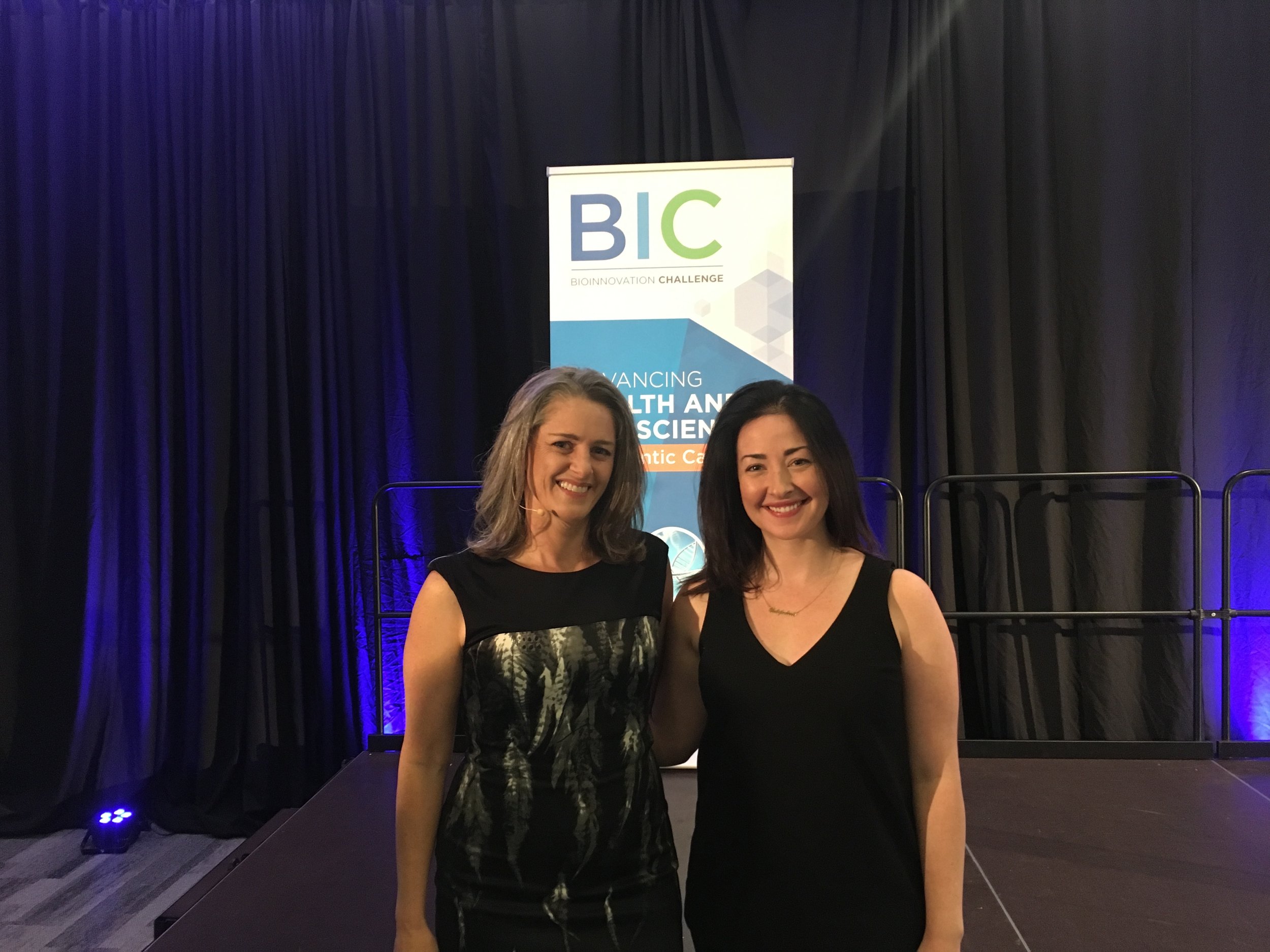 Two women standing on stage, smiling, in front of a banner that reads 'BIC BioInnovation Challenge' and 'Advancing Health and Science' at an indoor event.