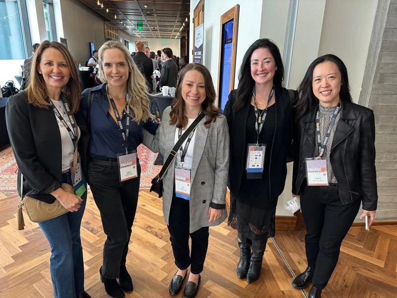Five women standing together at a professional conference, smiling at the camera, wearing conference badges, in an indoor setting with other attendees in the background.