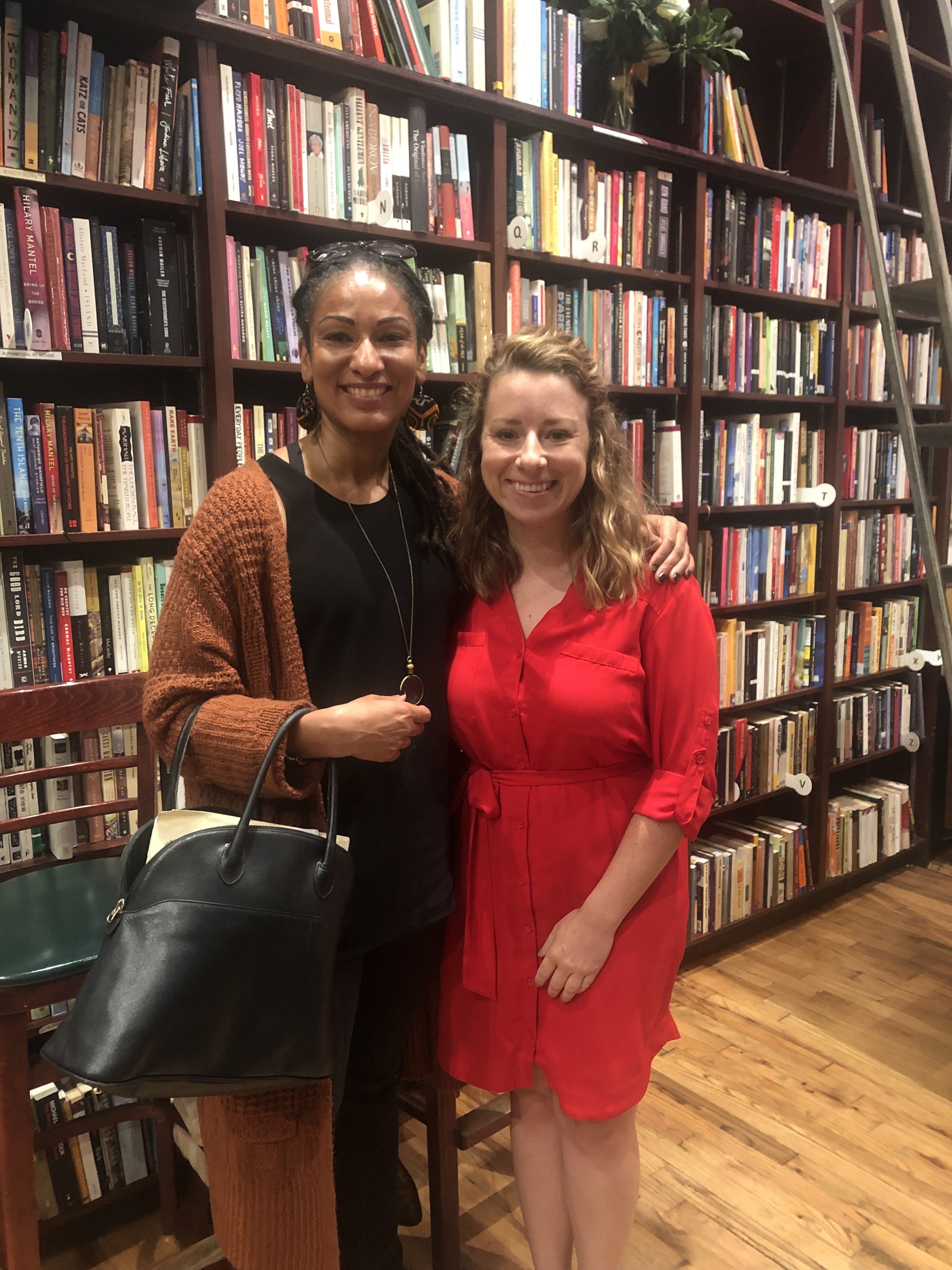 Two women standing closely together in front of bookshelves filled with colorful books in a cozy bookstore or library. The woman on the left is holding a black handbag and is wearing a black top with a brown cardigan, while the woman on the right is 