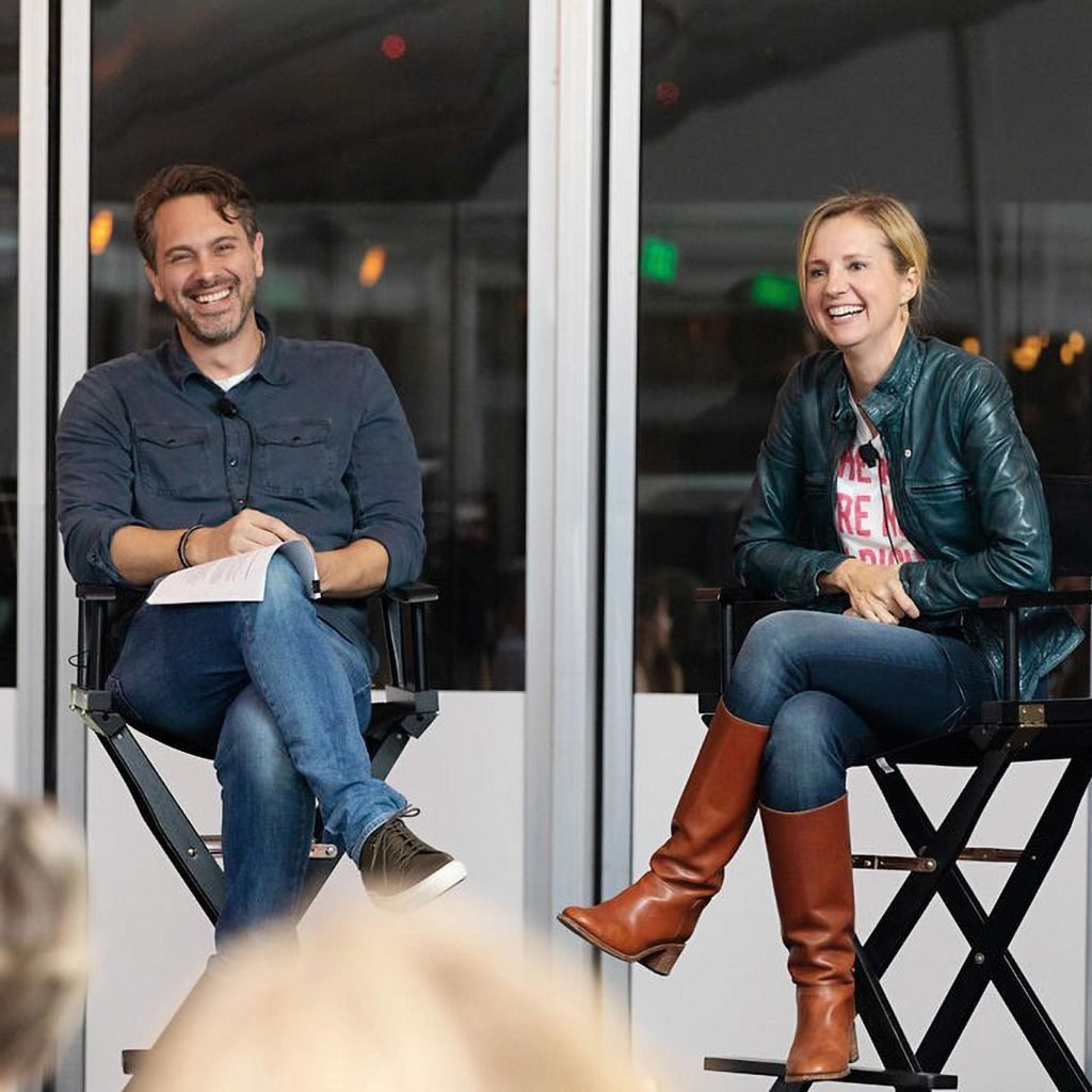 A man and woman sitting on director's chairs, laughing and engaged in conversation, in a glass-enclosed space at night.