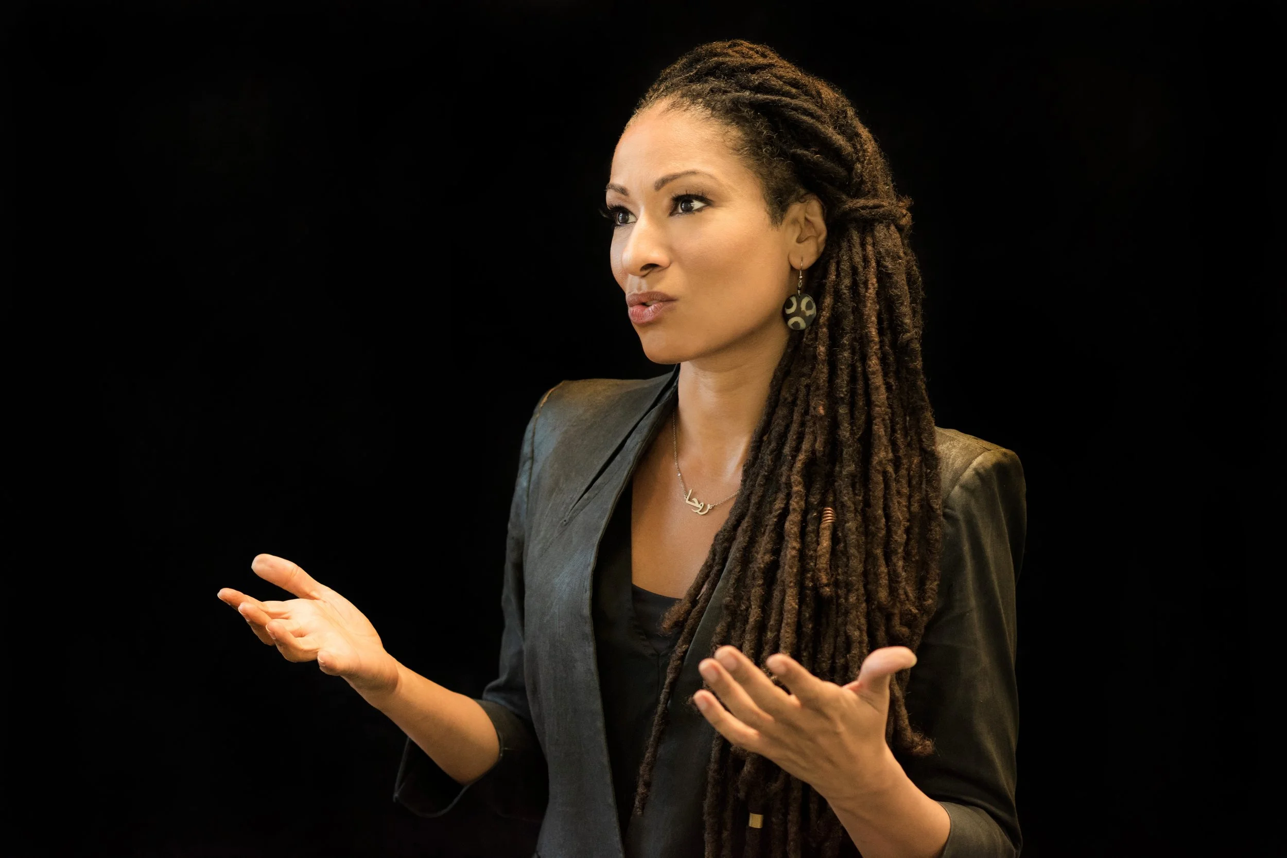 A woman with long dreadlocks, wearing a black blazer, speaking with her hands in front of a black background.