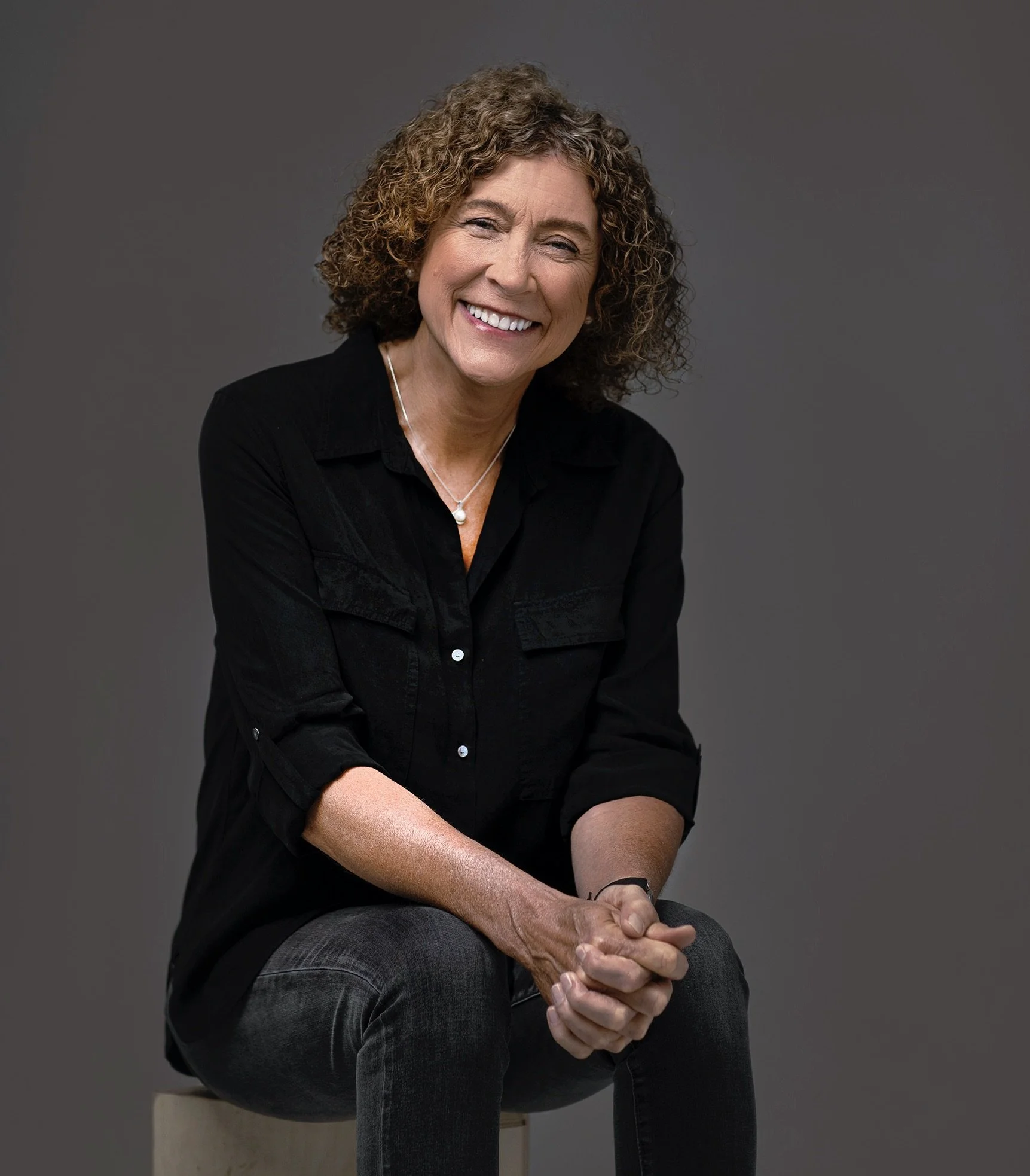 A smiling woman with curly hair, wearing a black shirt and gray jeans, sitting on a beige stool against a gray background.