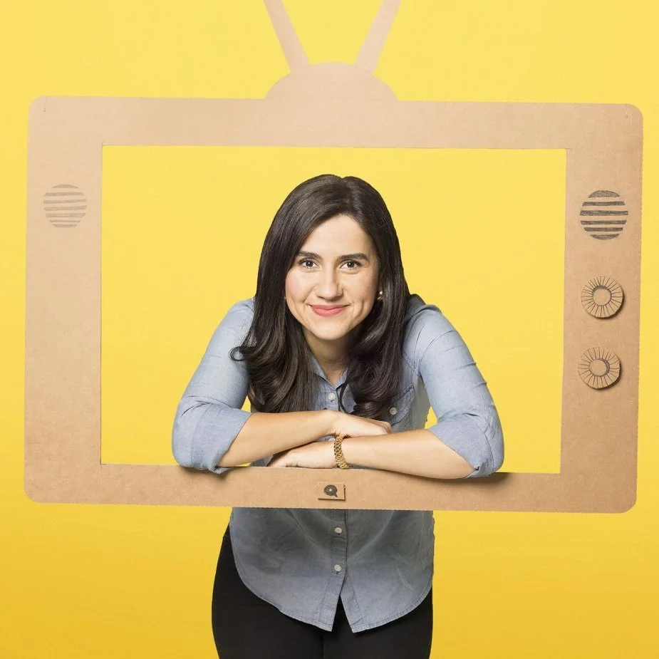 A woman with dark brown hair wearing a light blue shirt standing behind a cardboard cutout of a television frame, set against a yellow background.