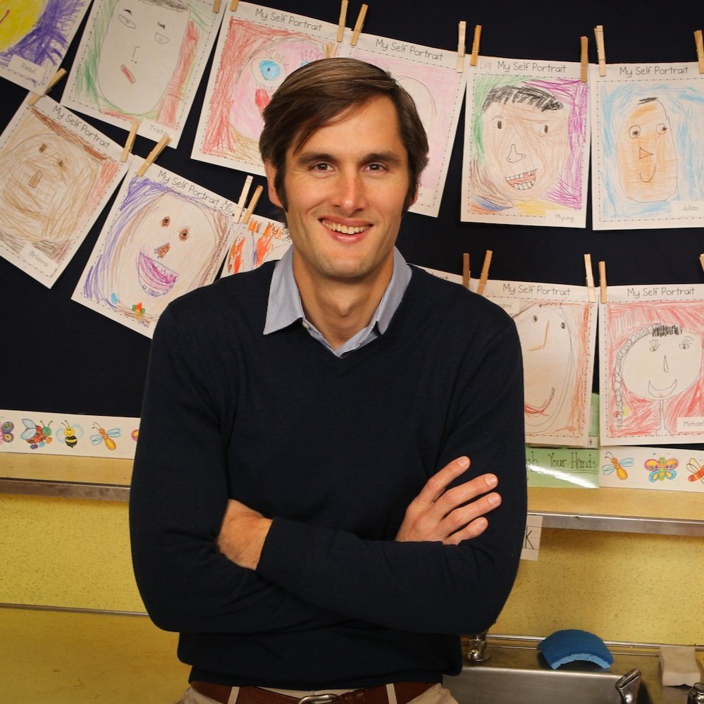 A man standing in front of a black display board with children's self-portrait drawings hanging on a string with clothespins, smiling with arms crossed.