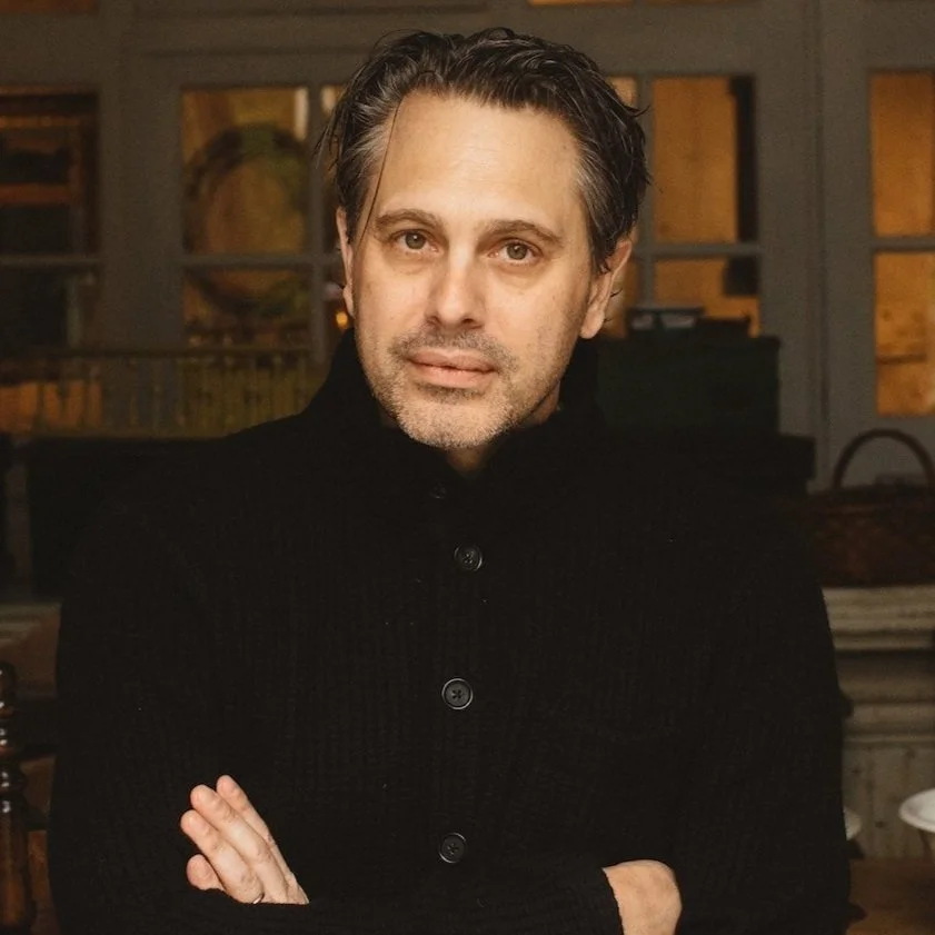 A man with brown hair and a beard, wearing a black sweater, standing indoors with arms crossed, in front of a window and a globe in the background.