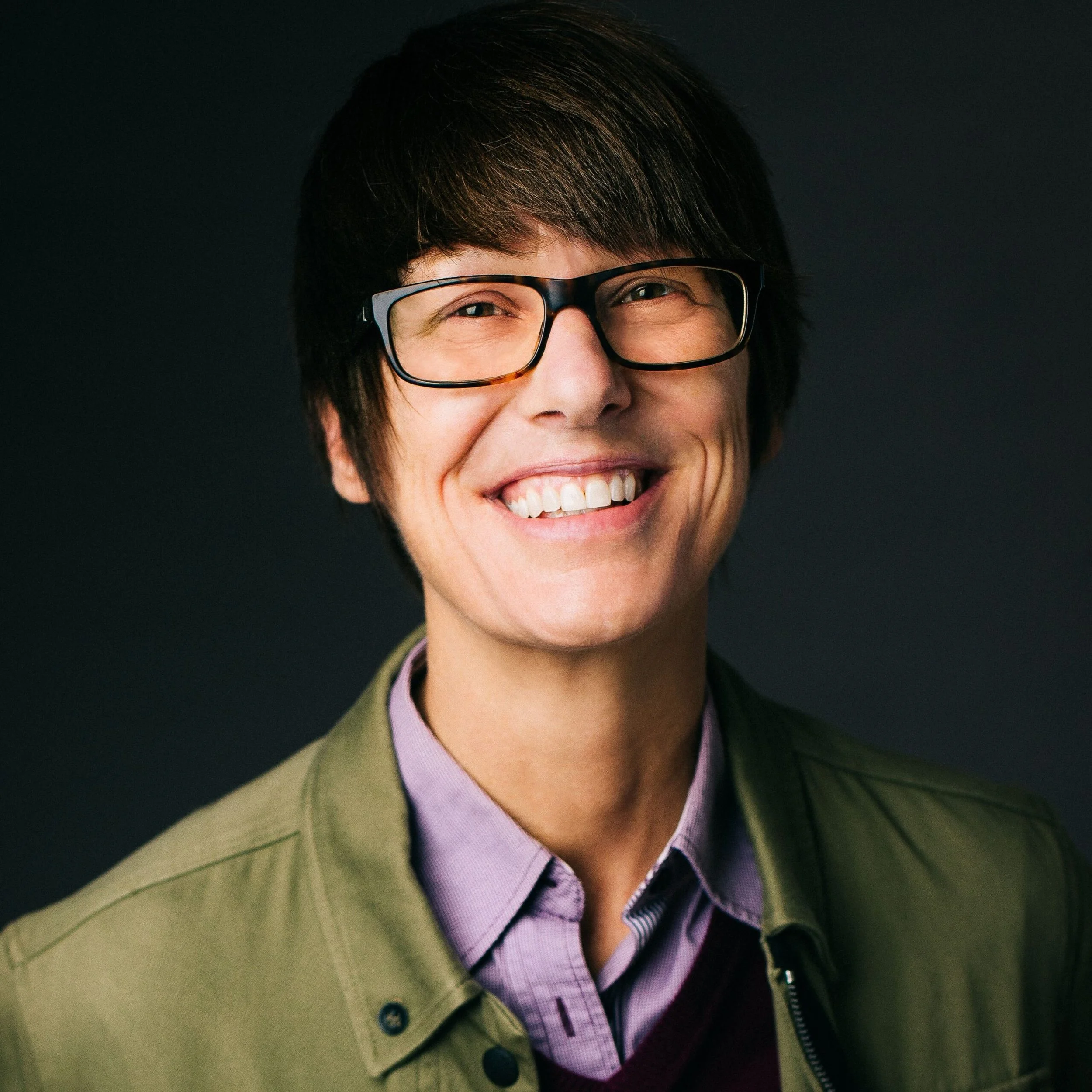 Portrait of a smiling woman with short dark hair wearing glasses, a purple collared shirt, and a green jacket against a dark background.