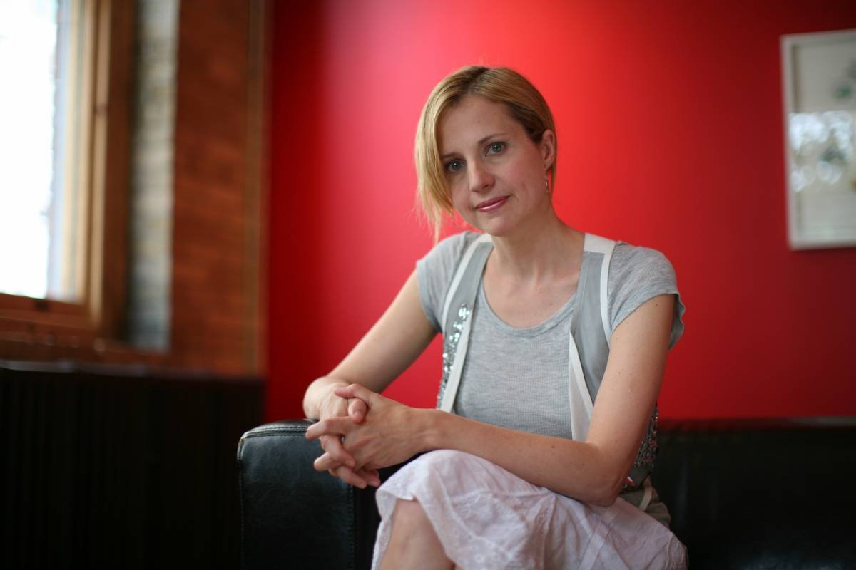 A woman with short red hair wearing a gray t-shirt sitting on a black chair, with a red wall behind her and a window on the left side.