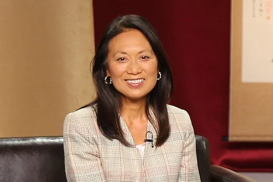 A smiling woman with shoulder-length dark hair, wearing a light-colored blazer, sitting in a chair indoors.