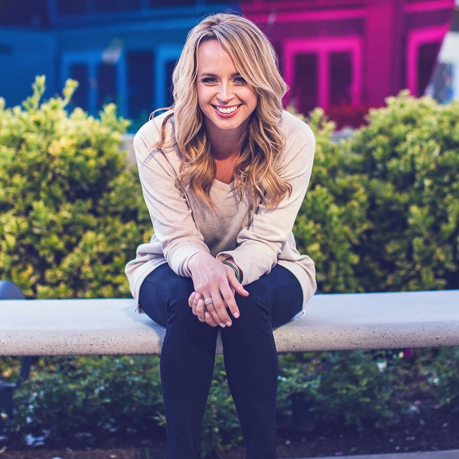 A smiling woman with blonde hair sitting on a concrete bench outdoors, with greenery and colorful buildings in the background.