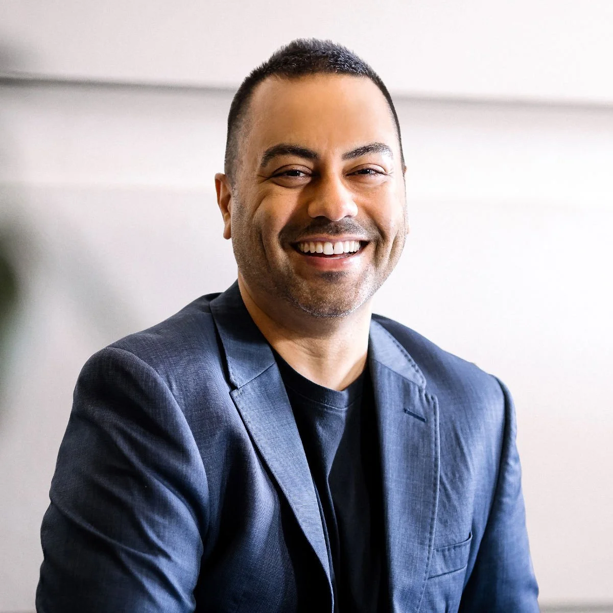 A smiling man with short dark hair, wearing a dark blue blazer over a black shirt, sitting indoors against a neutral background.