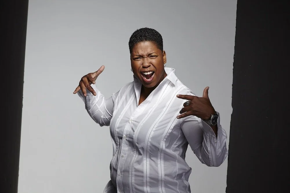 A woman with short hair making a rock and roll hand gesture, smiling widely with her eyes closed, wearing a white striped shirt, standing between black and gray backgrounds.