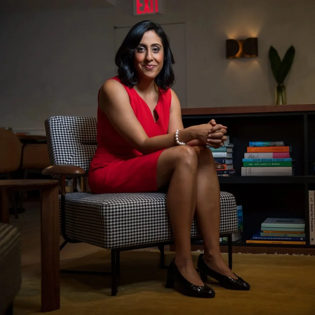 A woman in a red dress sitting on a houndstooth patterned chair in a room with bookshelves and warm lighting.
