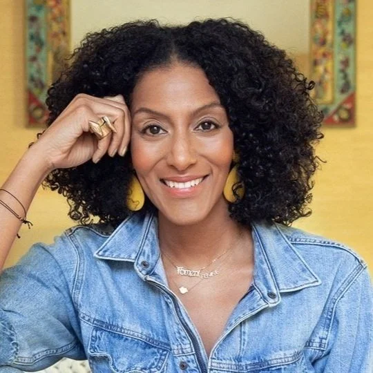 A woman with curly hair, smiling, wearing a denim jacket, yellow earrings, and a necklace with the name 'Tanya' on it.