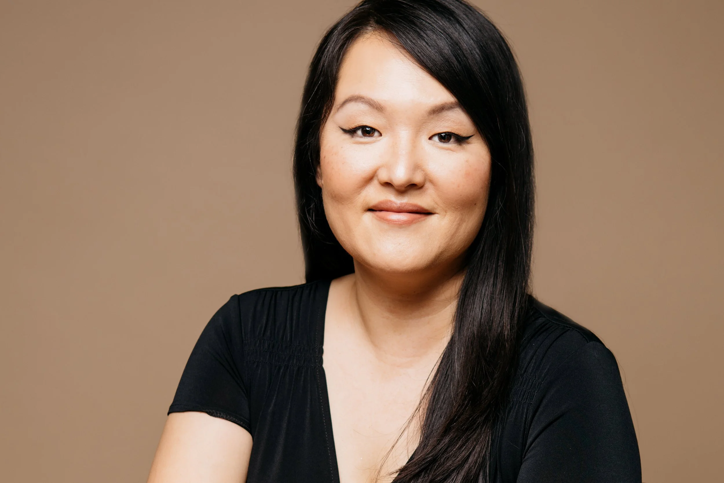 A woman with long black hair wearing a black top, smiling softly against a plain brown background.