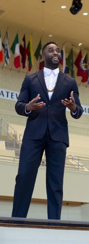 A man in a dark suit speaking on stage, with multiple international flags hanging in the background.