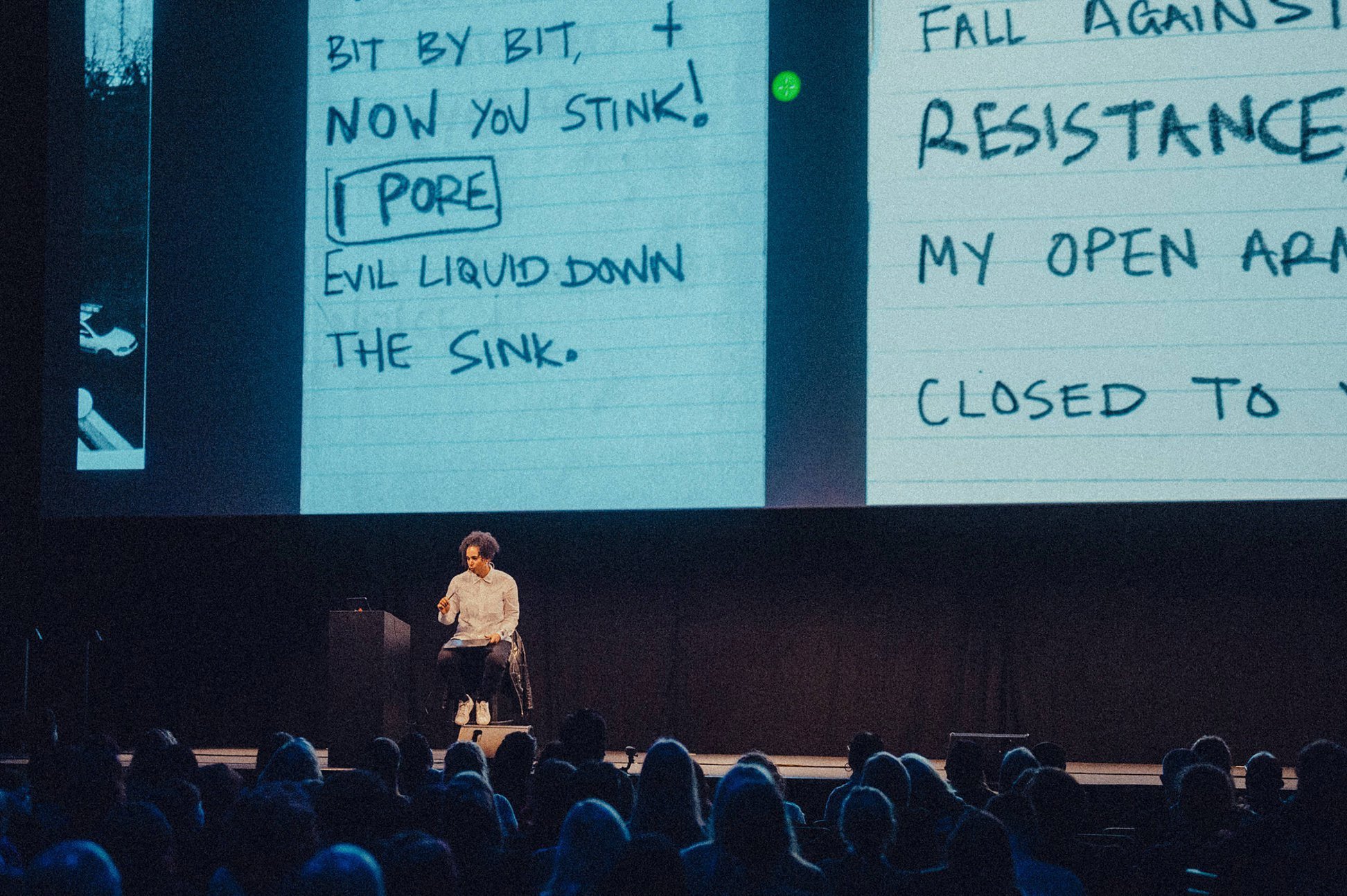 A woman is giving a presentation on stage with a large screen behind her showing handwritten notes about dealing with a sink leak. The notes mention evading an evil liquid and include a sketch of a pore.
