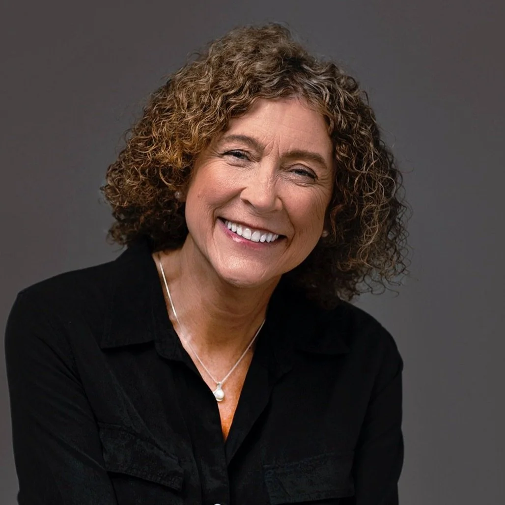 Portrait of a smiling middle-aged woman with curly short hair, wearing a black shirt and pearl necklace against a gray background.