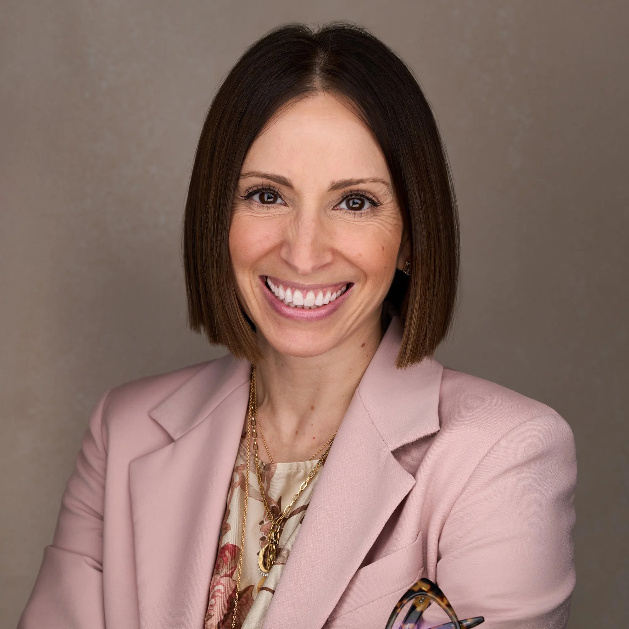 A woman with shoulder-length brown hair smiling outdoors, wearing a sleeveless green lace top and a pearl necklace, with a blurred green foliage background.