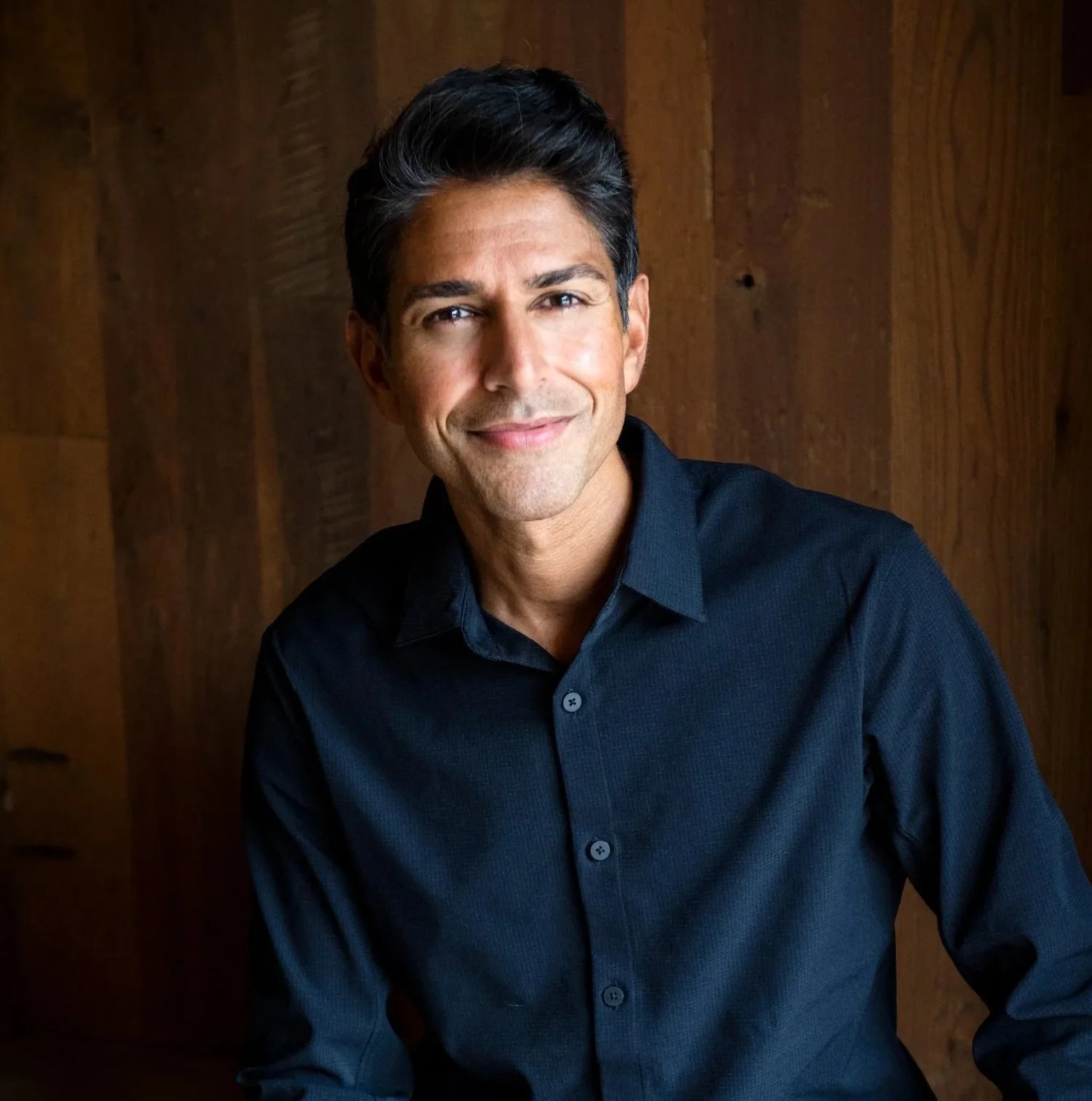 A man with dark hair wearing a dark blue shirt, smiling and looking at the camera, against a wooden background.