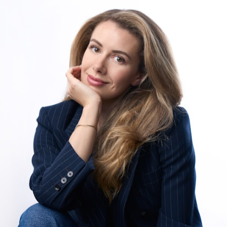 Portrait of a young woman with long, wavy hair, wearing a dark pinstripe blazer, smiling softly, with her hand resting on her cheek against a white background.