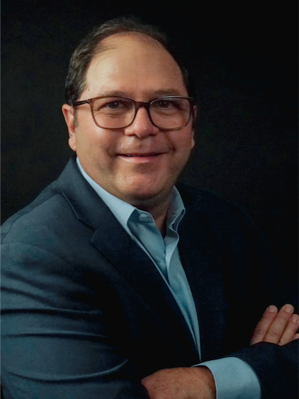 Portrait of a man with glasses in a dark background, wearing a blazer and light blue shirt, smiling with arms crossed.