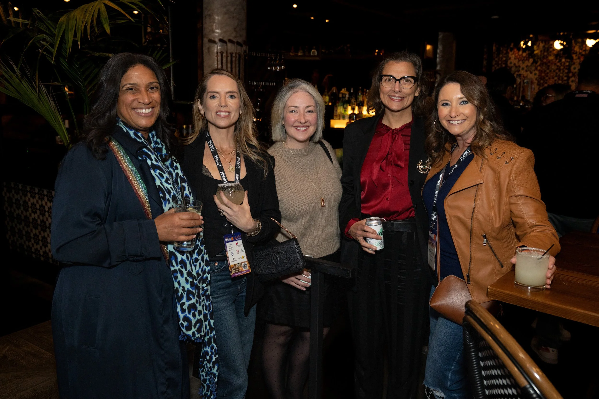 Group of five women smiling at social event in a bar or restaurant, holding drinks, with a bar and dim lighting in the background.