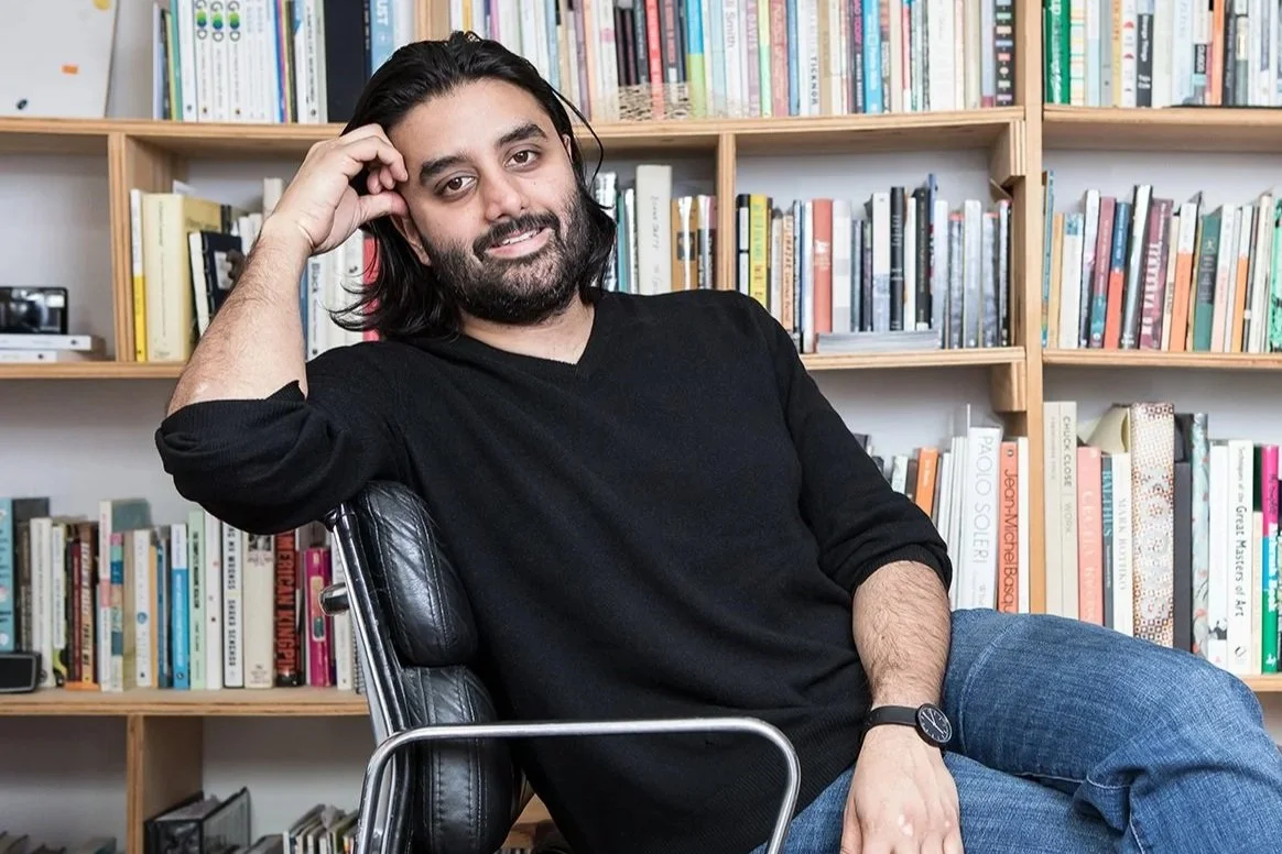 A man with long dark hair, a beard, and wearing a black long sleeve shirt, sitting in a black office chair with his right arm resting on the chair's armrest while leaning his head on his hand. He is in front of a wooden bookshelf filled with books.